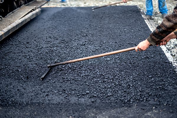 Worker leveling fresh asphalt on a road construction site, industrial buildings