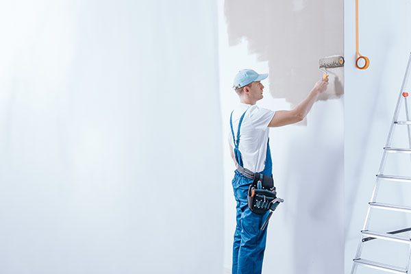 In the background painter in blue overalls painting white wall using roller