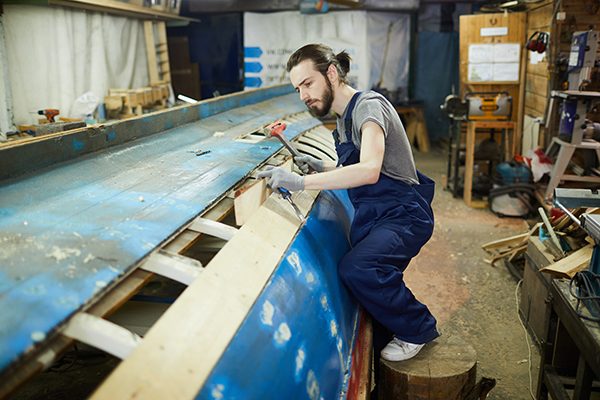 Professional shipbuilder in workwear hammering wooden board on vessel construction